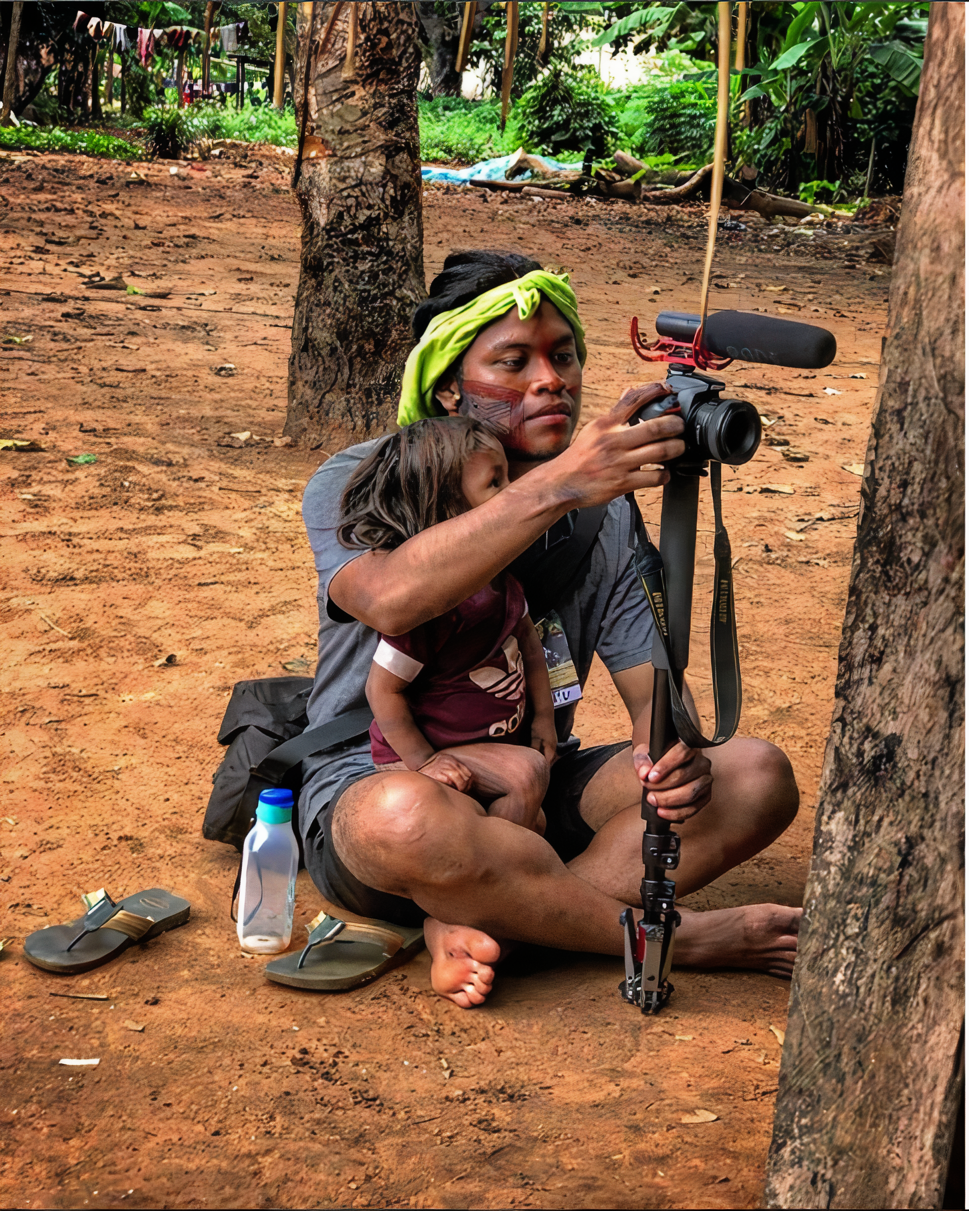 Cristian Wariu sentado, mexendo em uma câmera fotográfica, com uma criança pequena sentada em seu colo