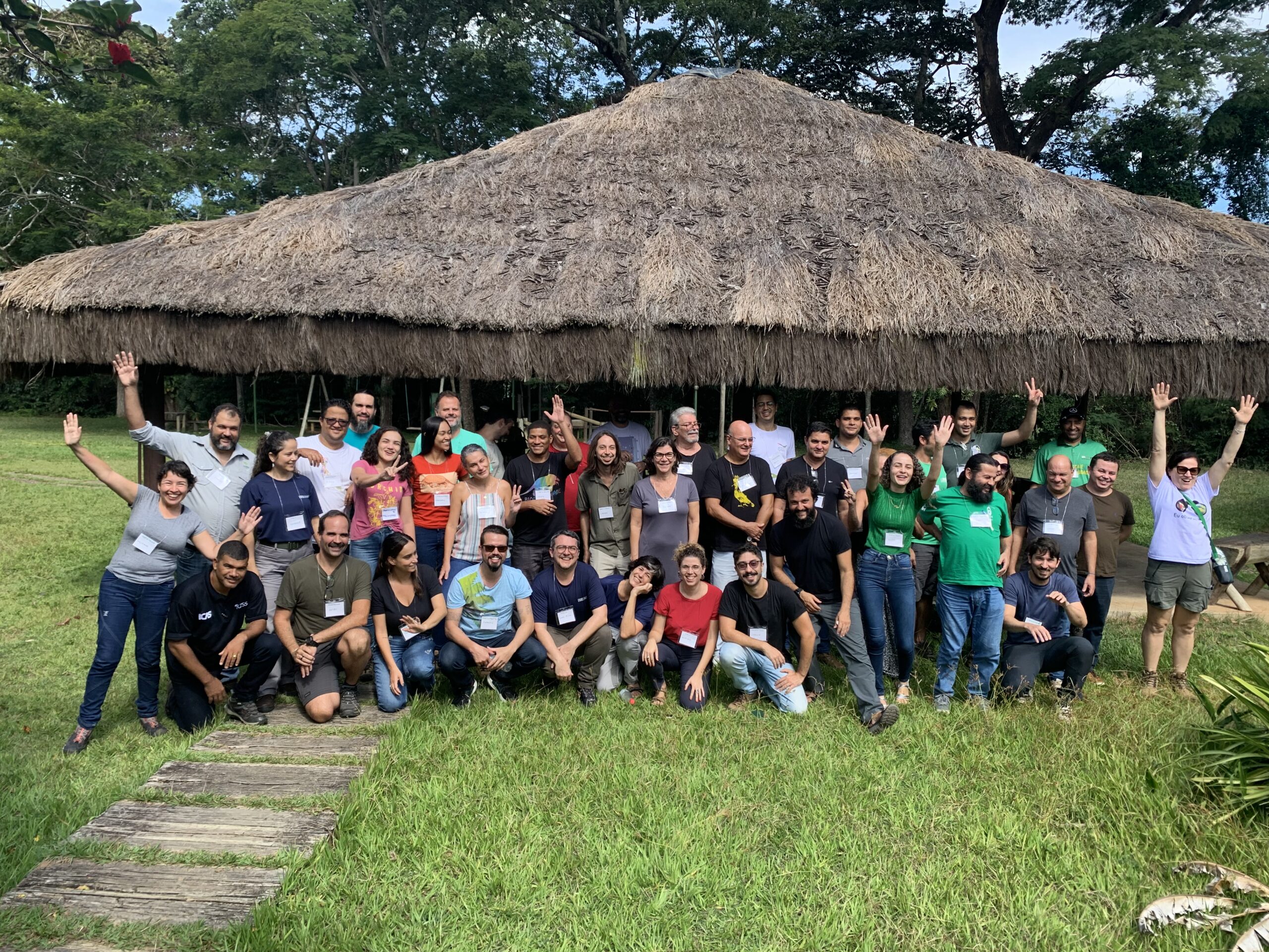 Foto com todos e todas que participaram do encontro de Revisão do plano de manejo do PERD. As pessoas estão do lado de fora, em cima de um gramado verde e atrás delas tem uma casa com telhado de um material aparentando palha. As pessoas estão de braços para cima e sorridentes.